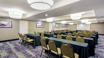 Un salón de conferencias con filas de mesas y sillas, ventanas amplias e iluminación en el techo.