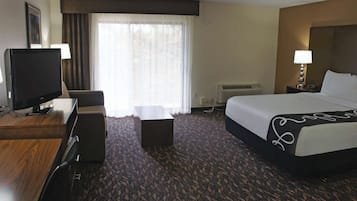 Et hotelværelse med en seng, et skrivebord med fjernsyn, en stol, et lille bord og et vindue med gardiner.