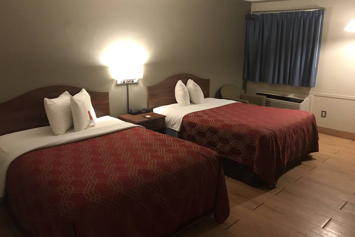 Habitación de hotel con dos camas, cada una con una colcha estampada roja y almohadas blancas.  Entre las camas hay una mesita de noche con una lámpara y un reloj. El piso es de madera y la ventana tiene cortinas azules.