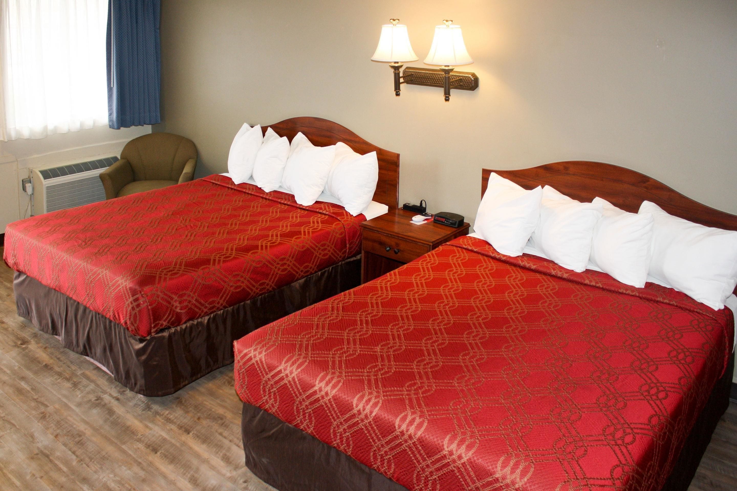 A hotel room with two beds, each with a red patterned bedspread and white pillows. There are two bedside tables with lamps, a chair, and a window with curtains.