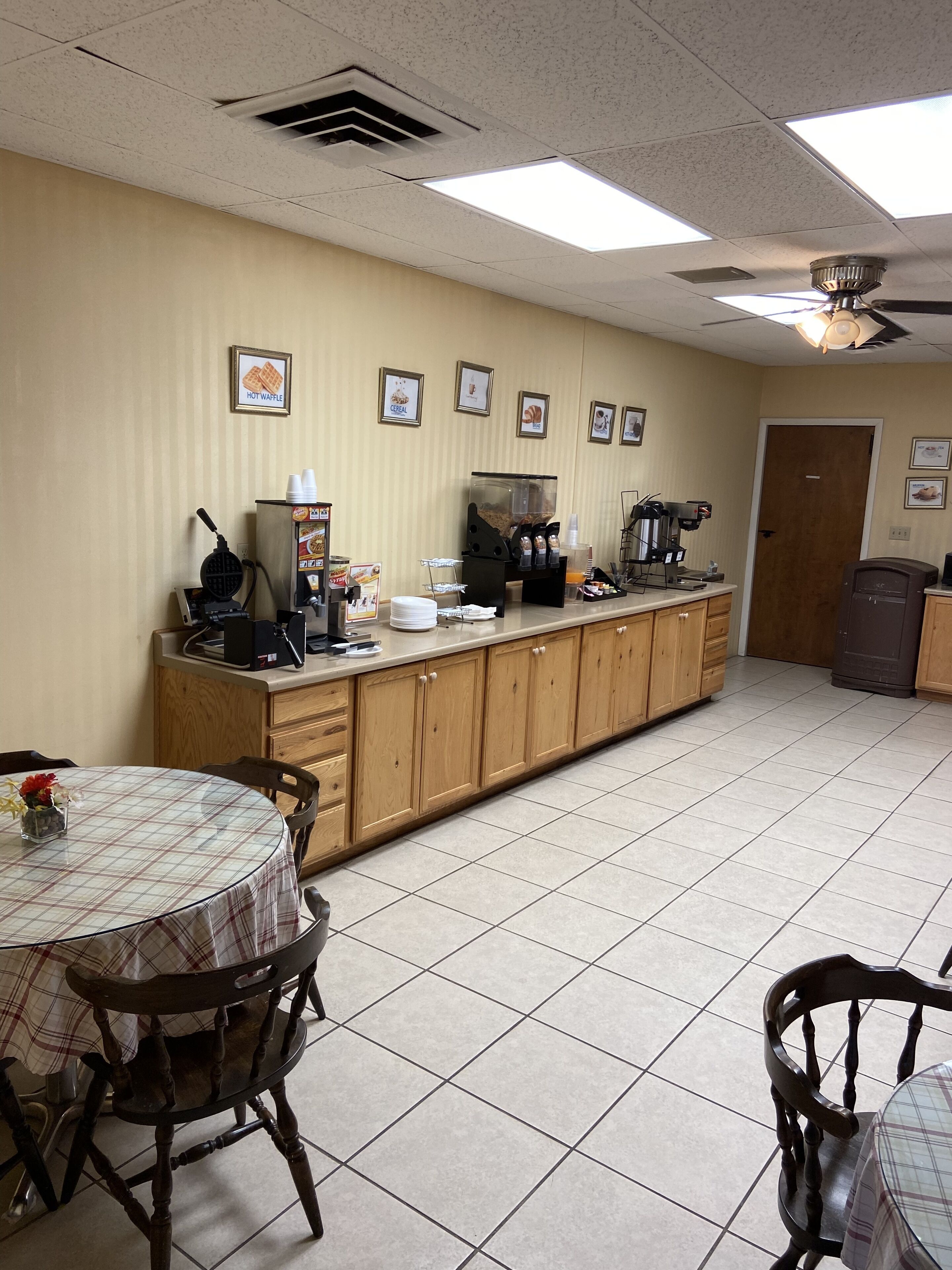 A clean, well-lit room with a long counter, a coffee machine, and a waffle maker. There are framed pictures on the wall and a door in the background.