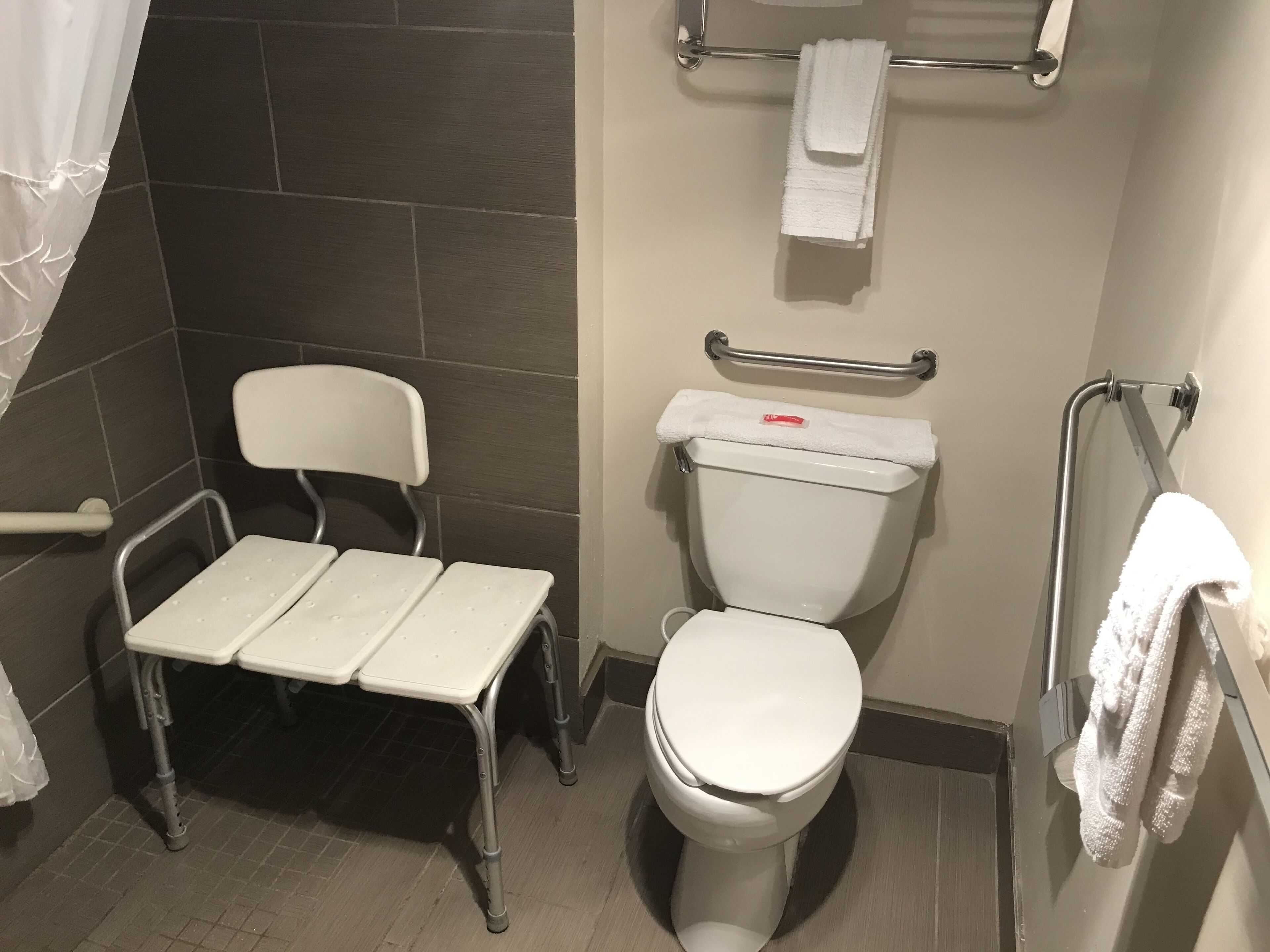A small, accessible bathroom with a white toilet, a white chair with armrests, a grab bar, and a towel rack.