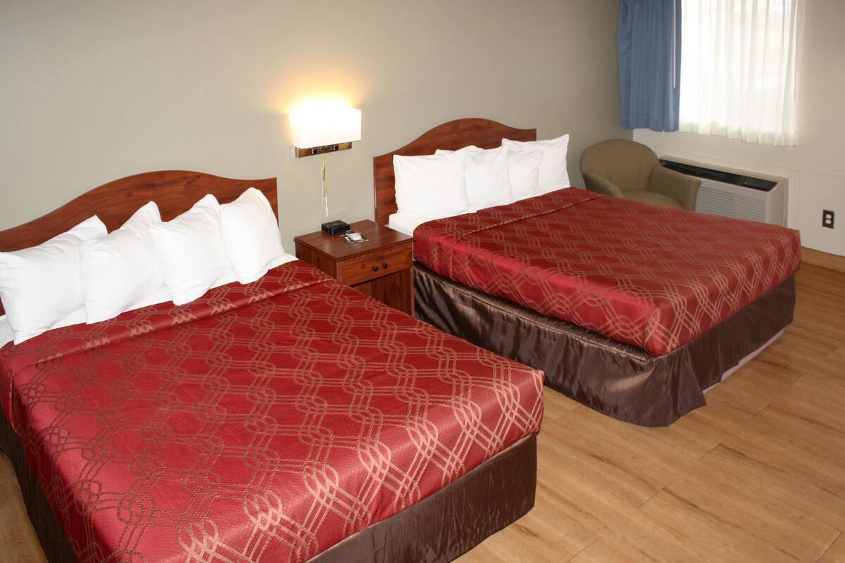 Una habitación de hotel con dos camas, una mesita de noche, una lámpara, una silla y una ventana con cortinas.