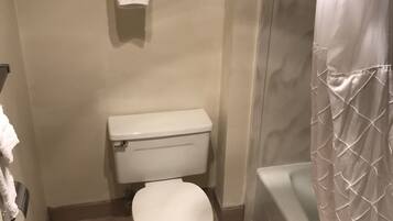 Une petite salle de bain avec des toilettes, une étagère où sont pliées des serviettes et un rideau de douche.