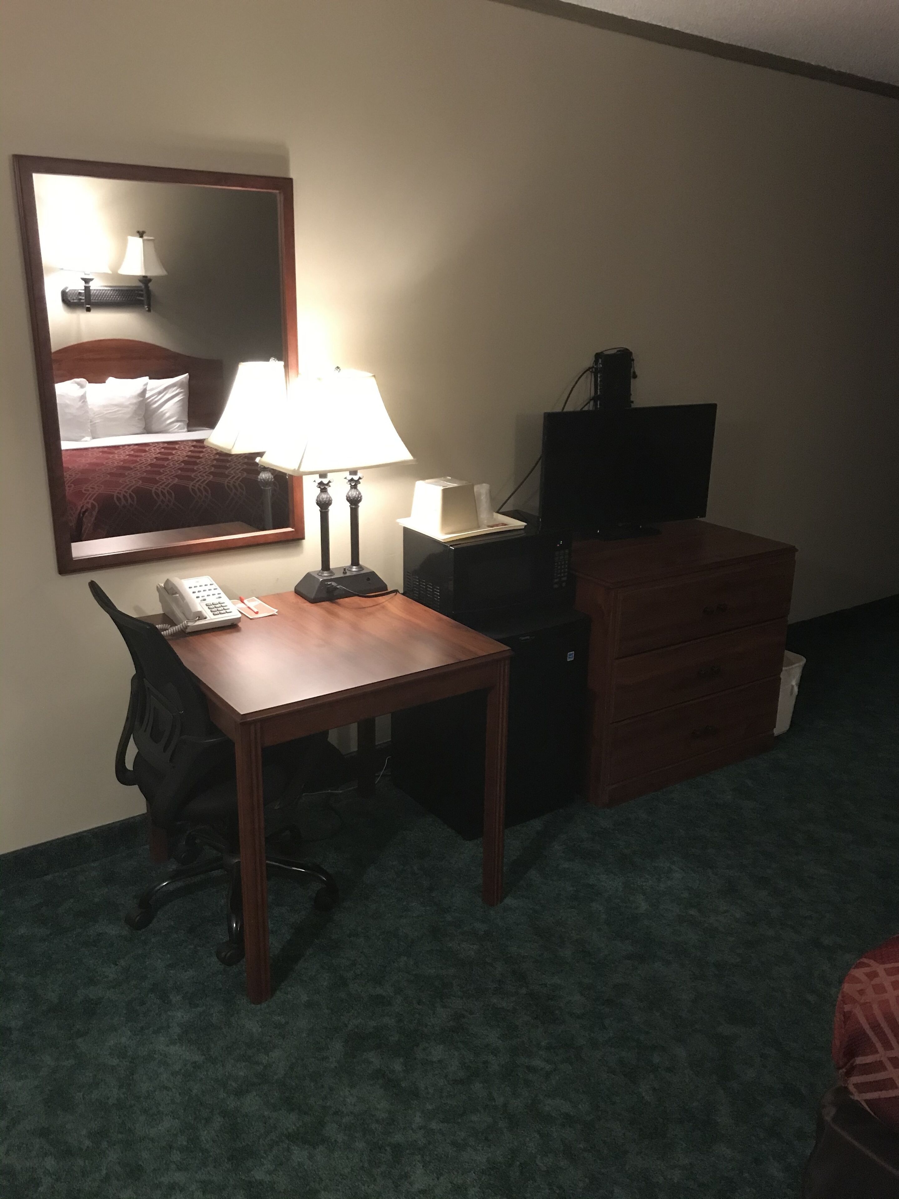Habitación de hotel con escritorio de madera, teléfono, lámpara, espejo, televisor y cómoda.
