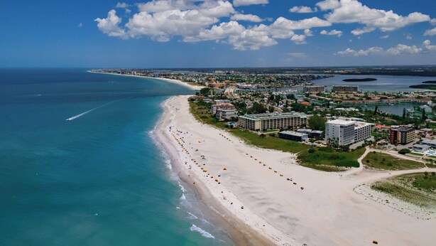 Luchtbeeld van een kuststad met een lang zandstrand, een jachthaven en diverse gebouwen, waaronder hotels.