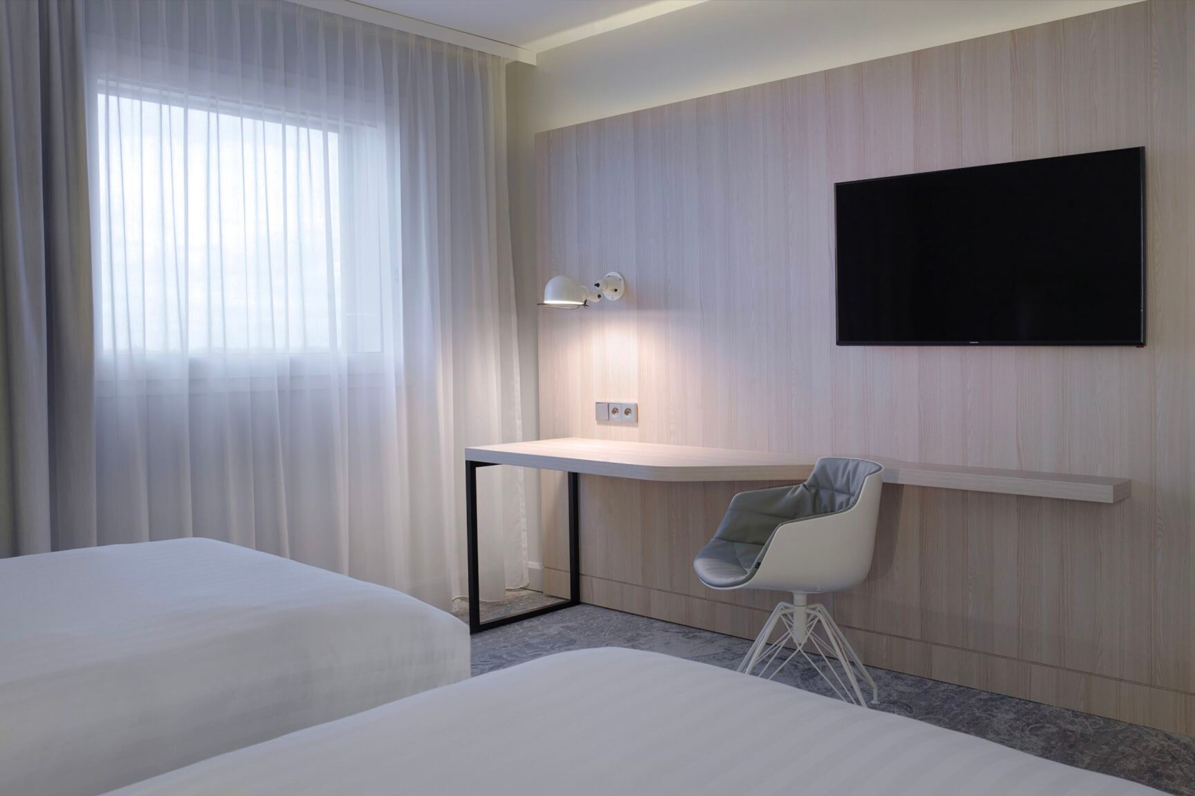 A hotel room with two beds, a desk with a chair, a mounted TV, and a window with curtains.