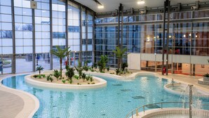 An indoor swimming pool with a circular design, surrounded by palm trees and a glass wall offering a view of the outside. - The Jangle Hotel - Paris - Charles de Gaulle - Airport (Le Mesnil-Amelot)