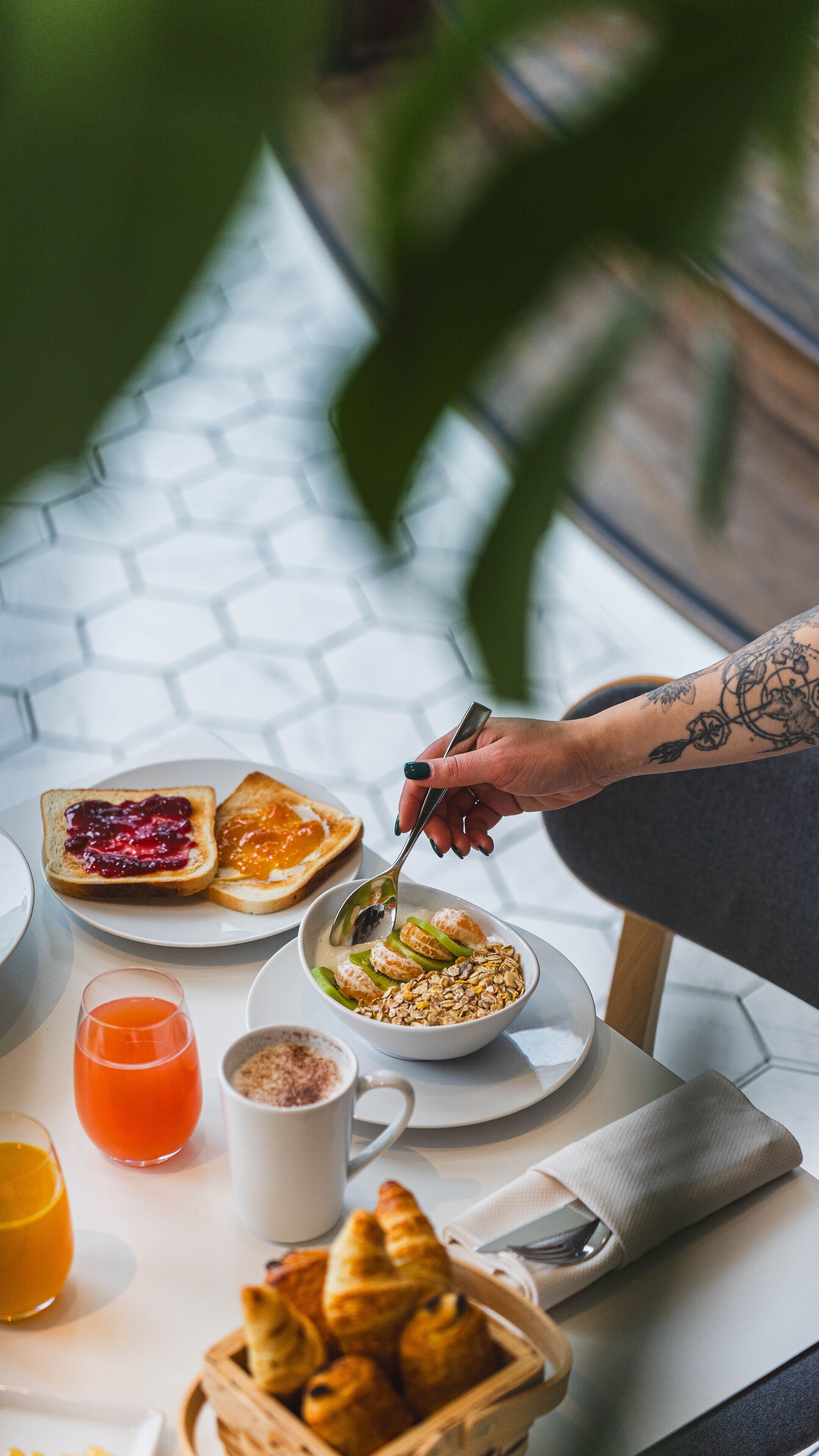A breakfast setting with toast, jam, orange juice, a bowl of cereal with fruit, and croissants on a table.