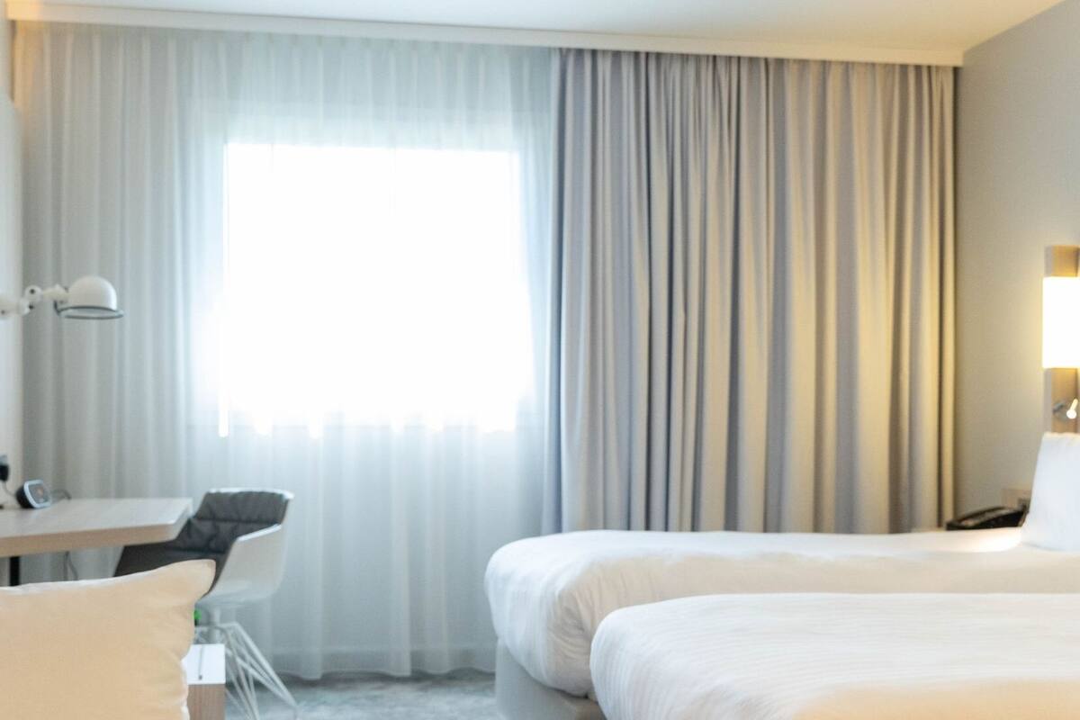A hotel room with a bed, a desk, and a chair. There is a window with curtains and a lamp on the wall.