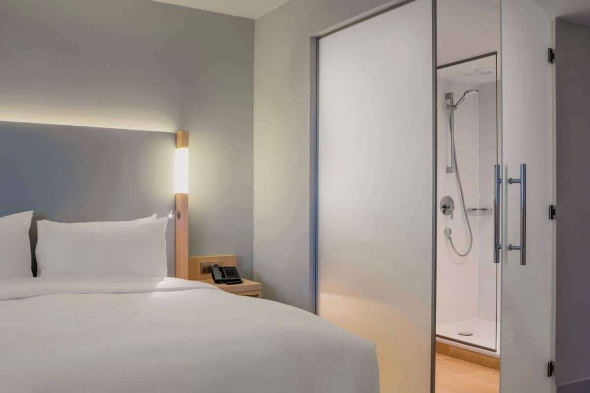 A modern hotel room with a large bed, bedside table with a phone, and a glass-enclosed shower area.