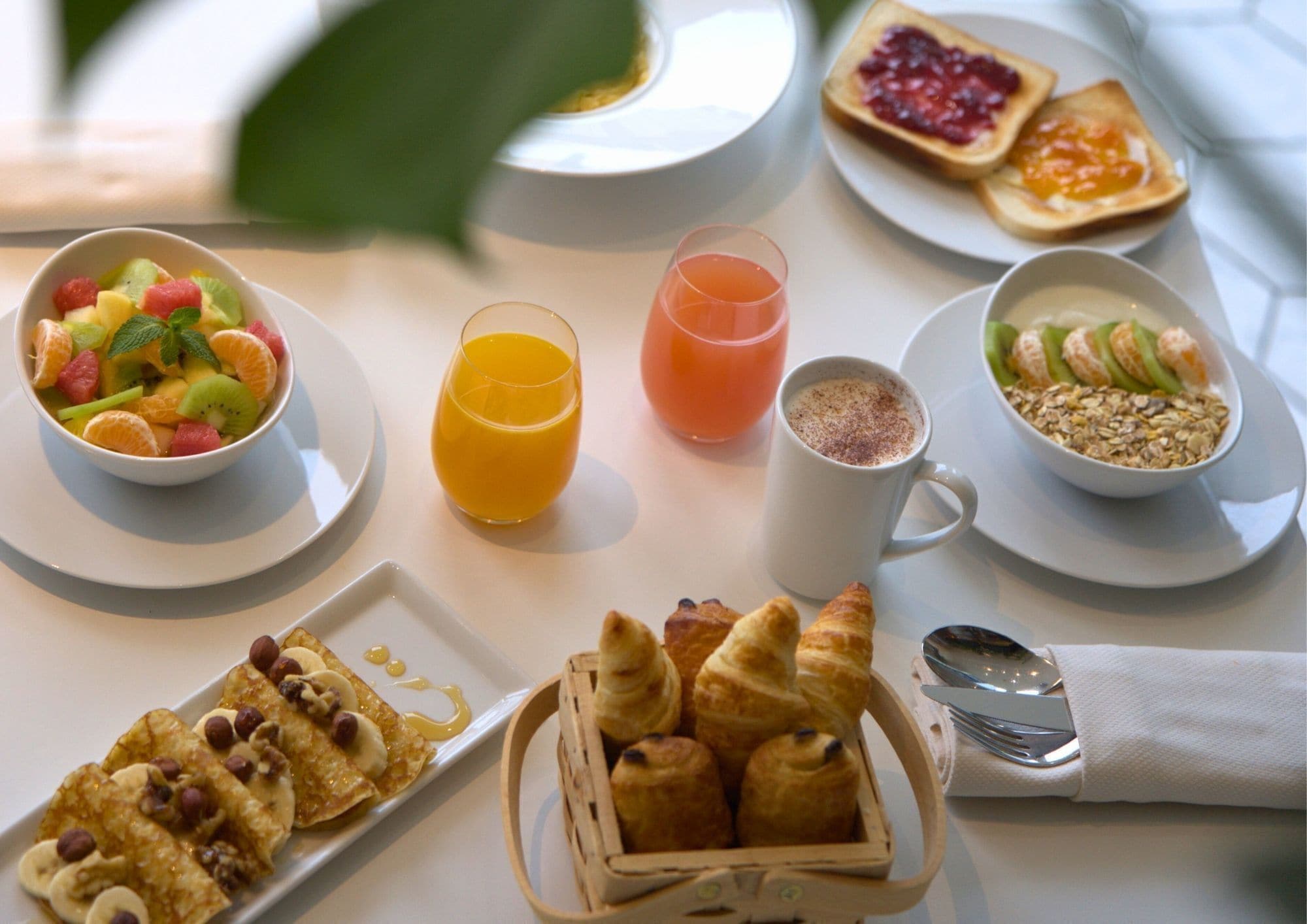 Un desayuno con un bol de ensalada de frutas, dos vasos de zumo de naranja, una taza de café, un plato de croissants y un plato de tostadas con mermelada.