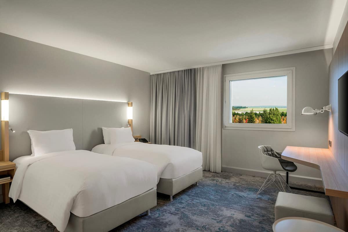A hotel room with two beds, a desk, a chair, and a large window with a view of greenery.
