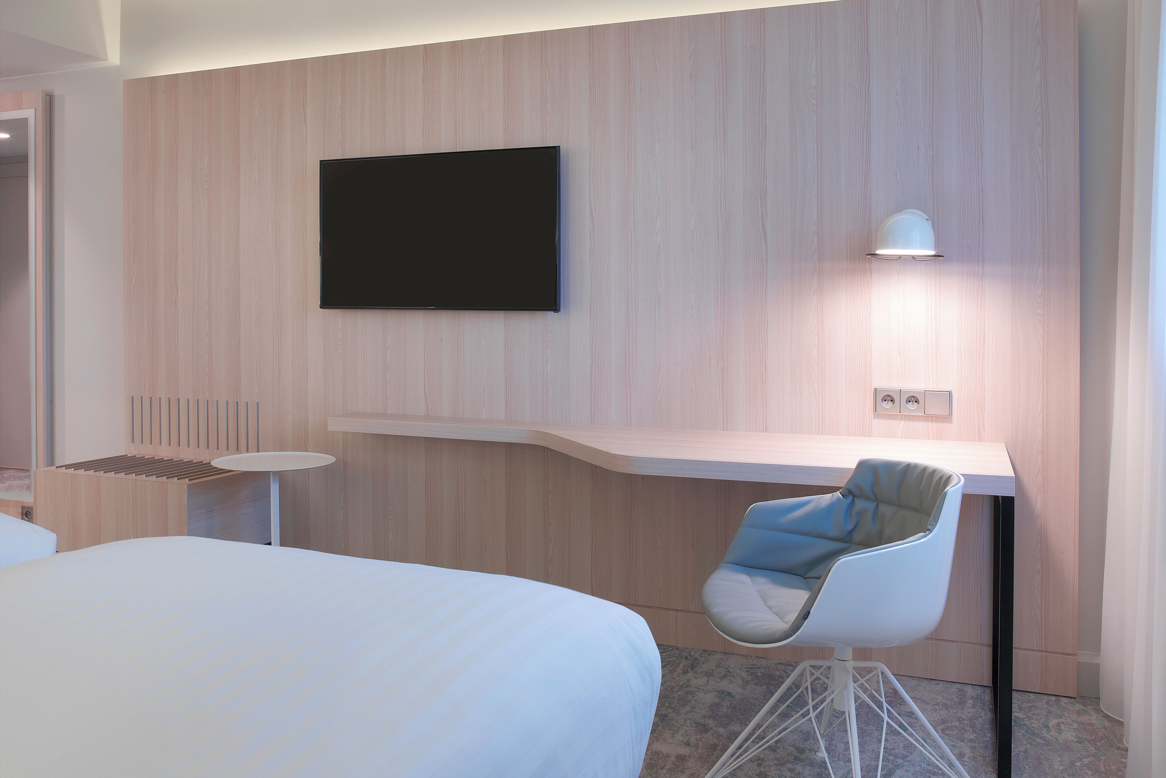 A modern hotel room with a bed, a desk with a chair, a flat-screen TV, and a wall-mounted lamp.