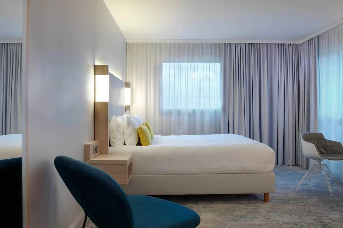 A modern hotel room with a large bed, a bedside table with a lamp, a chair, and a large window with curtains.