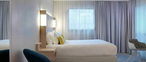 A modern hotel room with a large bed, a bedside table with a lamp, a chair, and a large window with curtains.