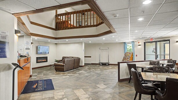 A hotel lobby with a herringbone floor, a reception desk, a television, a fireplace, a staircase, and a dining area with leather chairs. - Baymont by Wyndham Greensburg (Greensburg)