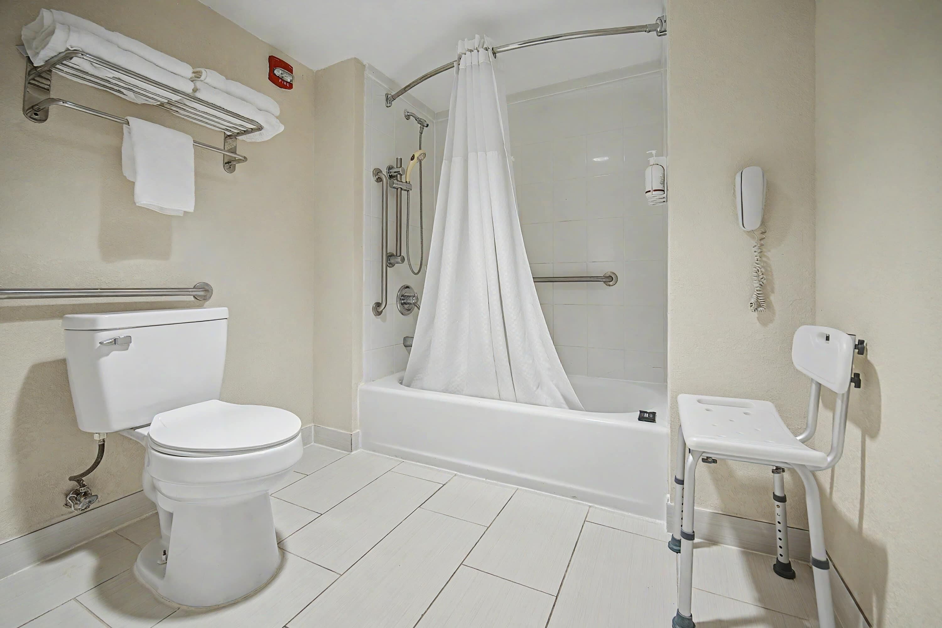 A bathroom with a toilet, a shower with a curtain, a grab bar, a white chair, and a phone on the wall.