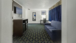 A hotel room with a patterned carpet, a sofa, a desk with a chair, a TV, a mirror, and a framed picture on the wall. - Baymont by Wyndham Greensburg (Greensburg)