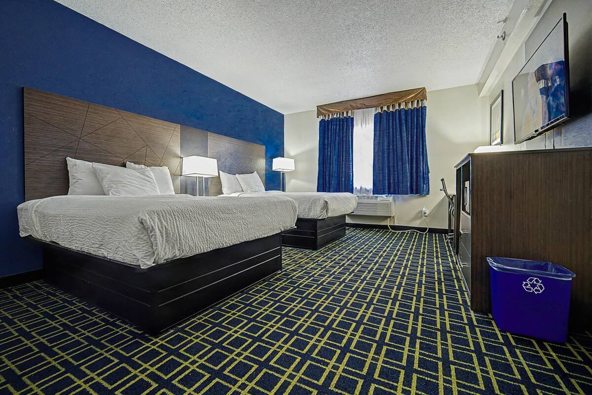 Une chambre d’hôtel avec deux lits, un bureau, une poubelle et une grande fenêtre avec des rideaux.