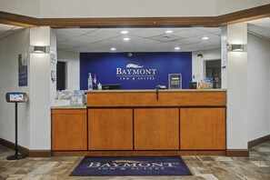 A reception desk with a blue wall and the Baymont Inn & Suites logo. There are informational posters on the wall, a reception counter, and a digital kiosk to the left. - Baymont by Wyndham Greensburg (Greensburg)