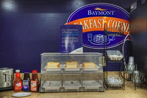 A breakfast counter with a Baymont sign, condiment bottles, a toaster, and a display case with food items. - Baymont by Wyndham Greensburg (Greensburg)