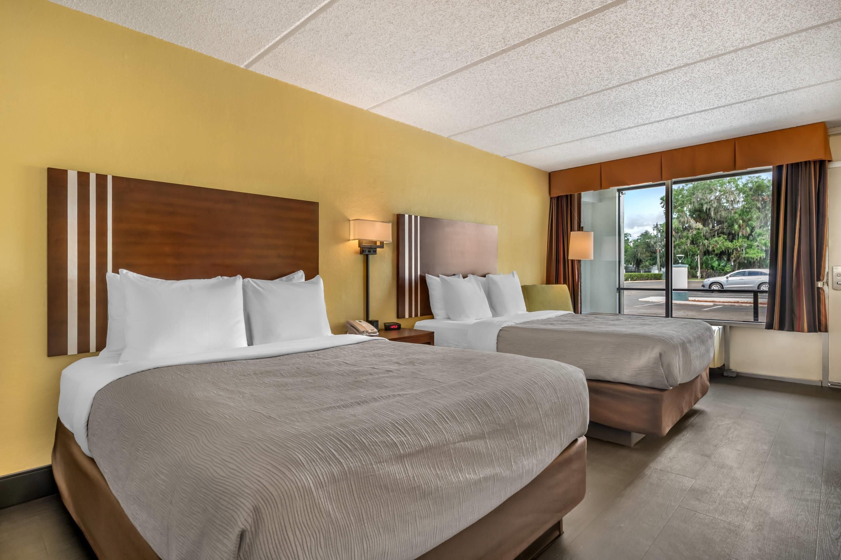 A hotel room with two beds, a nightstand, a lamp, and a window with a view of the outside.