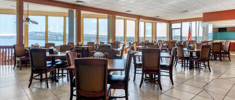 A spacious dining area with multiple tables and chairs, large windows offering views of a bridge and water, and an American flag on the wall.