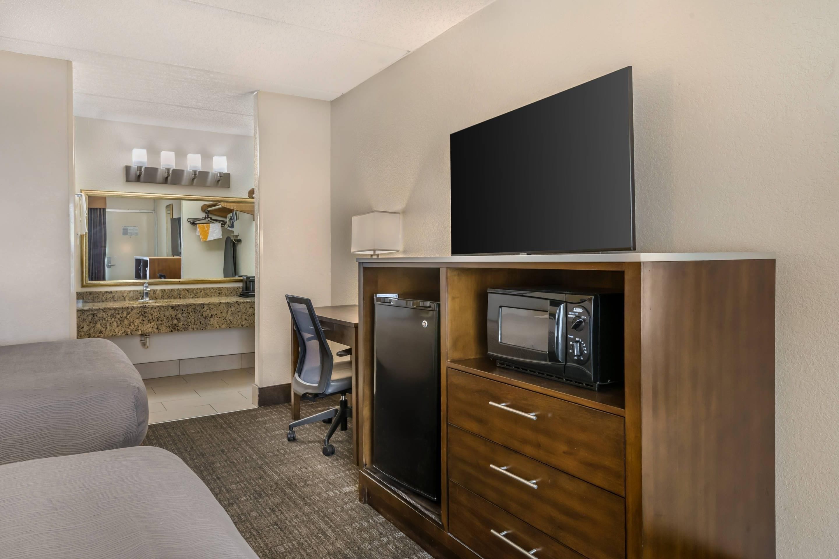 A hotel room with a bed, a desk, a chair, a microwave, a refrigerator, a flat-screen TV, and a mirror with lights above it.