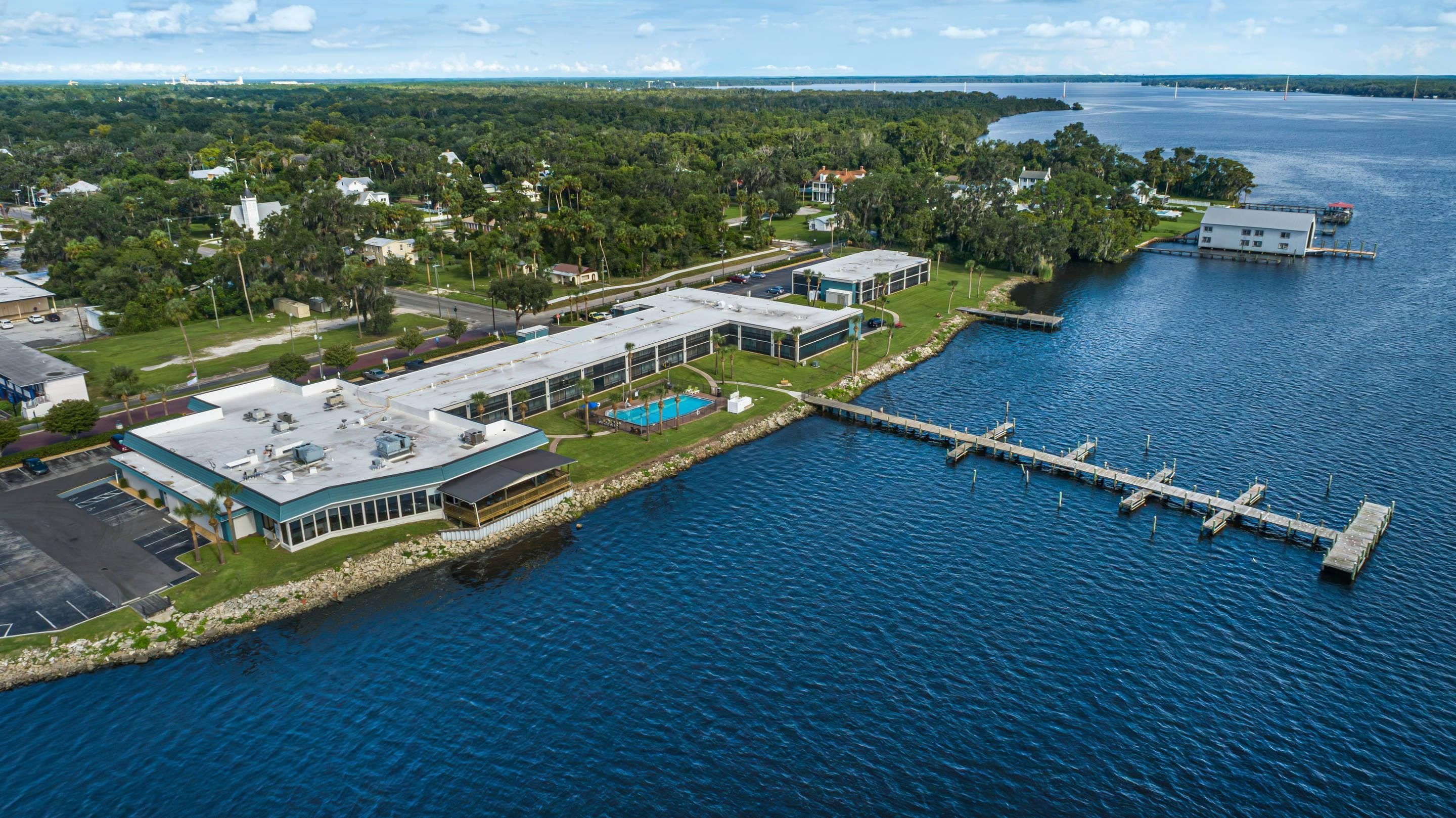 An aerial view of a waterfront property with a dock extending into a body of water, surrounded by greenery and residential buildings.
