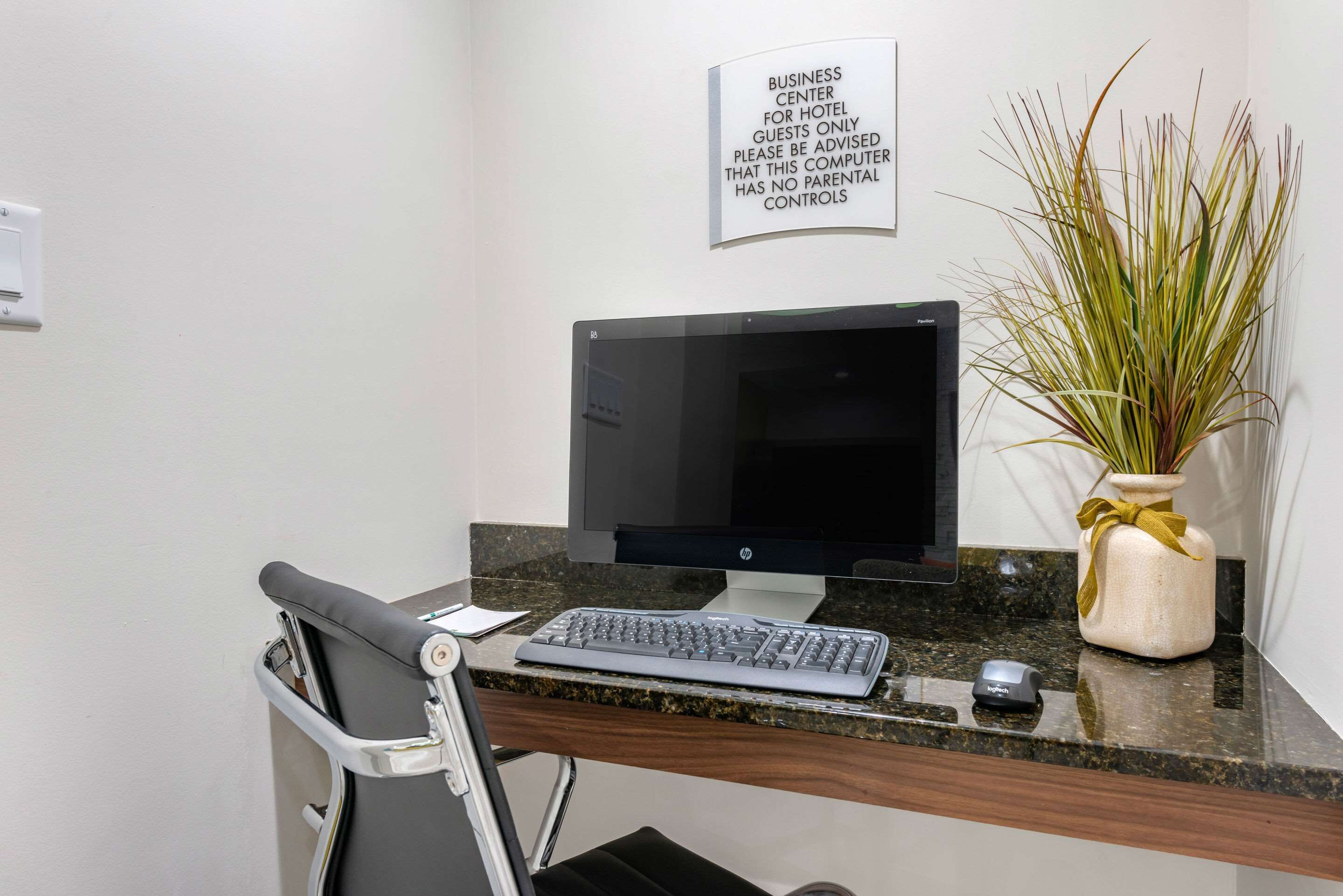 A hotel business center desk with a computer, keyboard, and mouse. A sign above the desk advises guests that the computer has no parental controls.
