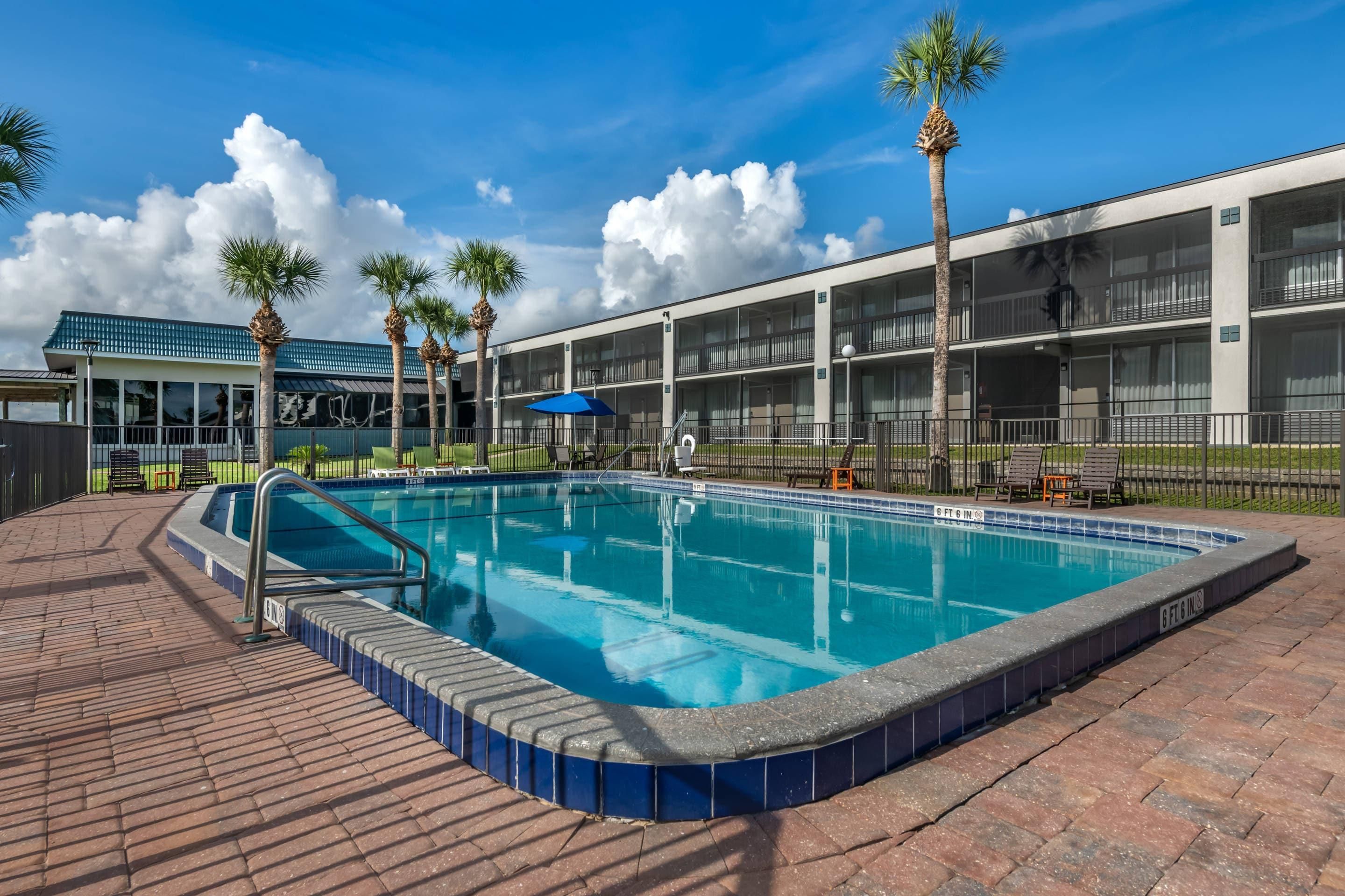 A swimming pool with a 6 ft depth, surrounded by a brick deck, palm trees, and a building with balconies.