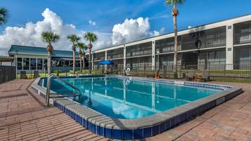 A swimming pool with a 6 ft depth, surrounded by a brick deck, palm trees, and a building with balconies.