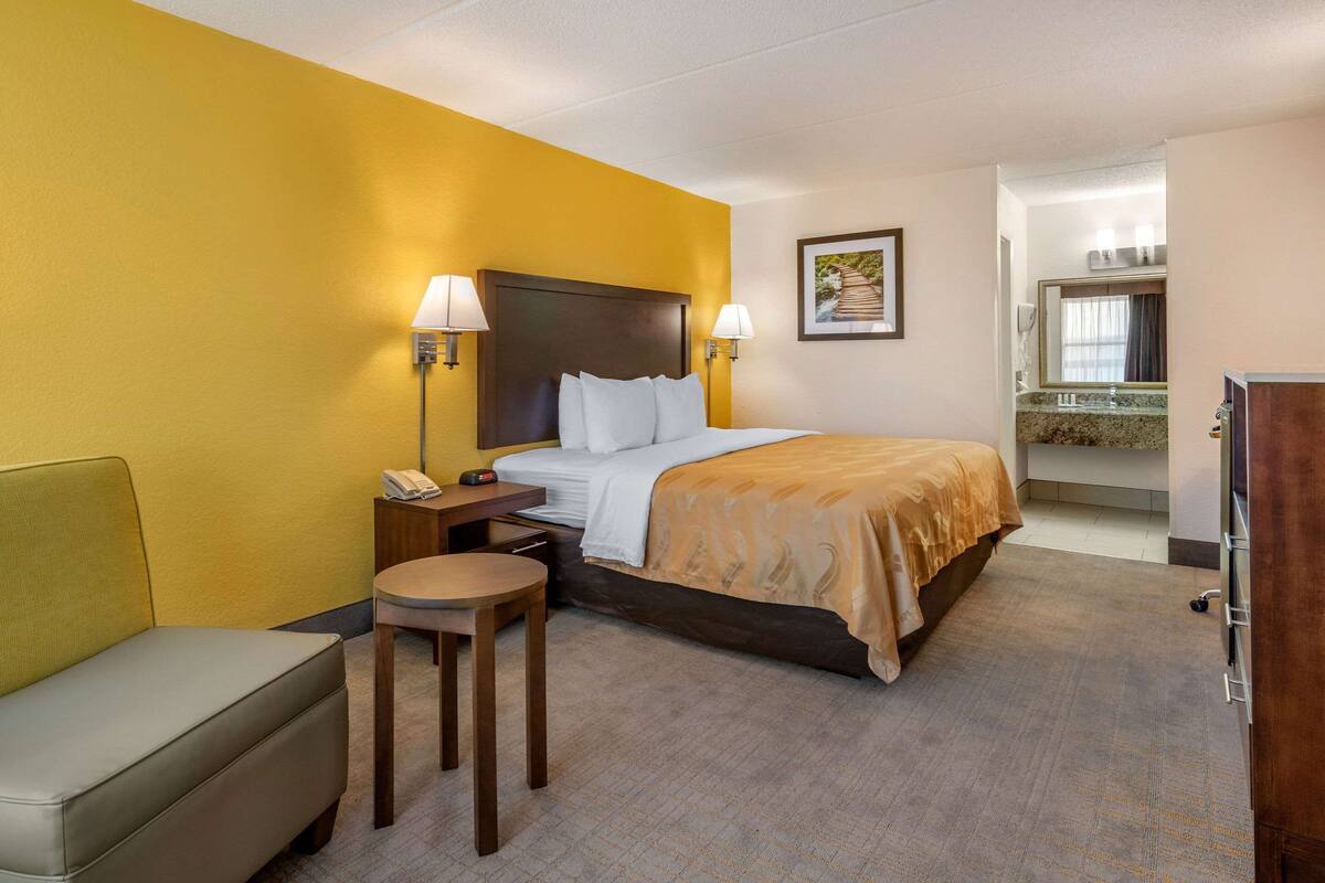 Habitación de hotel con cama, mesitas de noche, una silla, un escritorio y un baño visible a través de una puerta abierta.