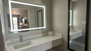 A bathroom with a white countertop, a sink, a mirror with a light, and a small trash bin.