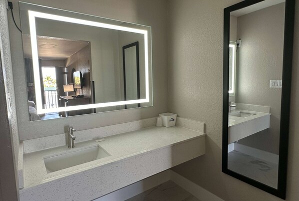 A bathroom with a white countertop, a sink, a mirror with a light, and a small trash bin. - Days Inn by Wyndham Fort Myers (Fort Myers)
