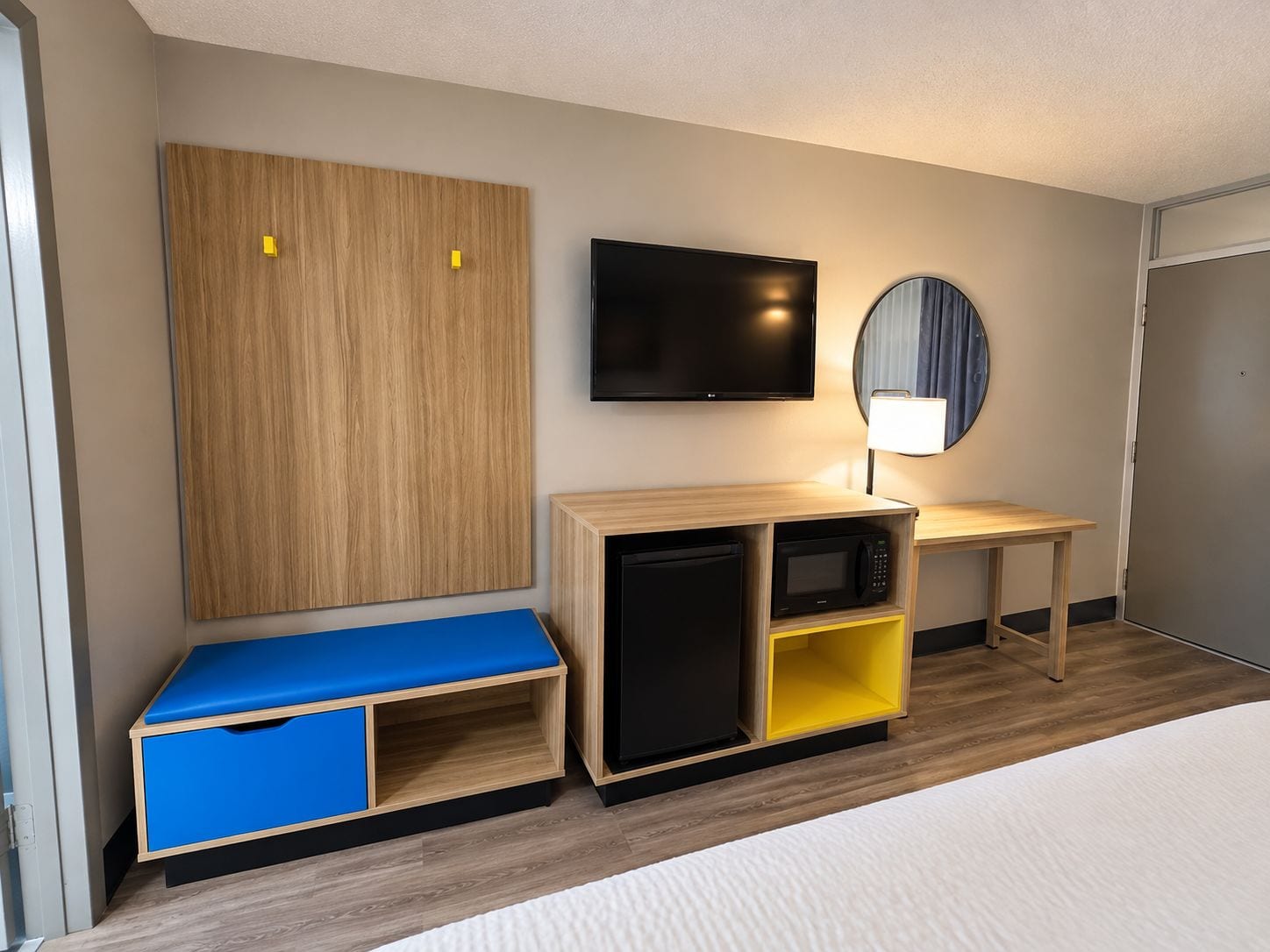 A hotel room with a bed, a TV, a desk with a lamp, a wardrobe, and a small kitchenette.