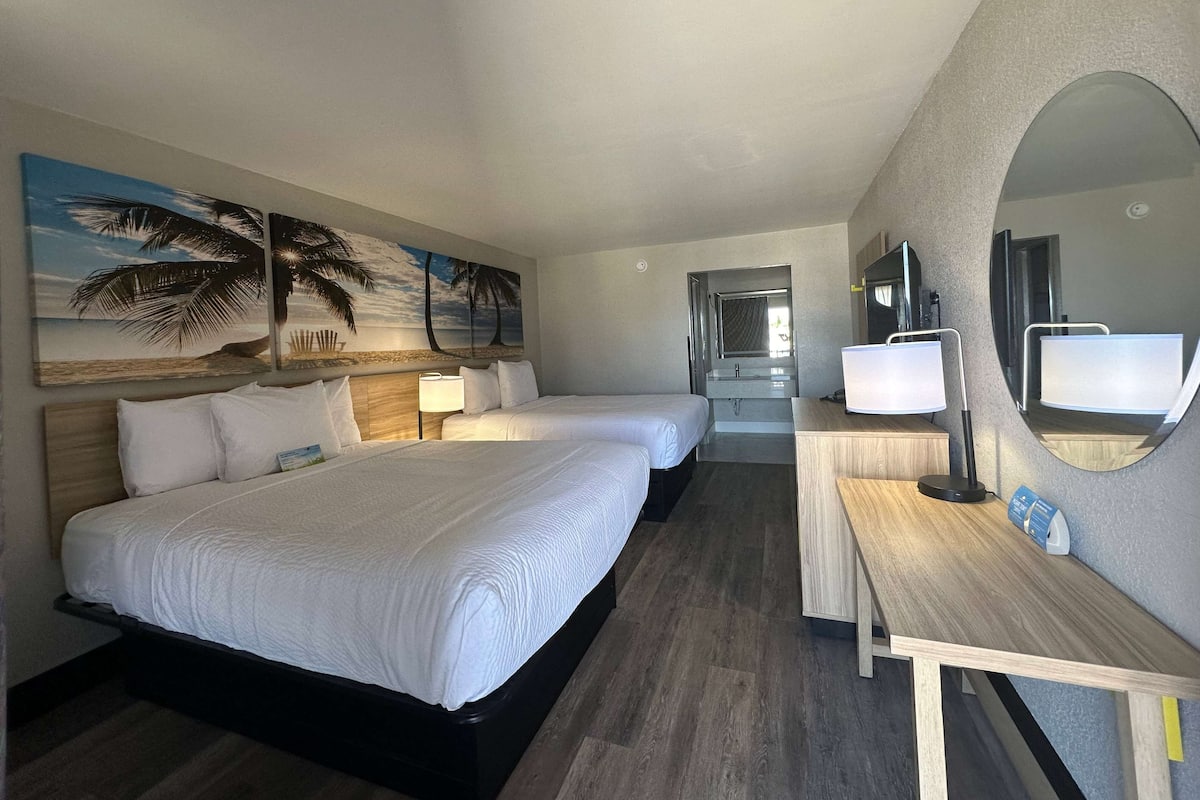 A hotel room with two beds, a desk, a mirror, and a painting of a beach scene.