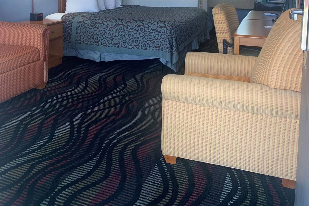 Habitación de hotel con cama, dos sillas, escritorio, lámpara, un cuadro y alfombra estampada.
