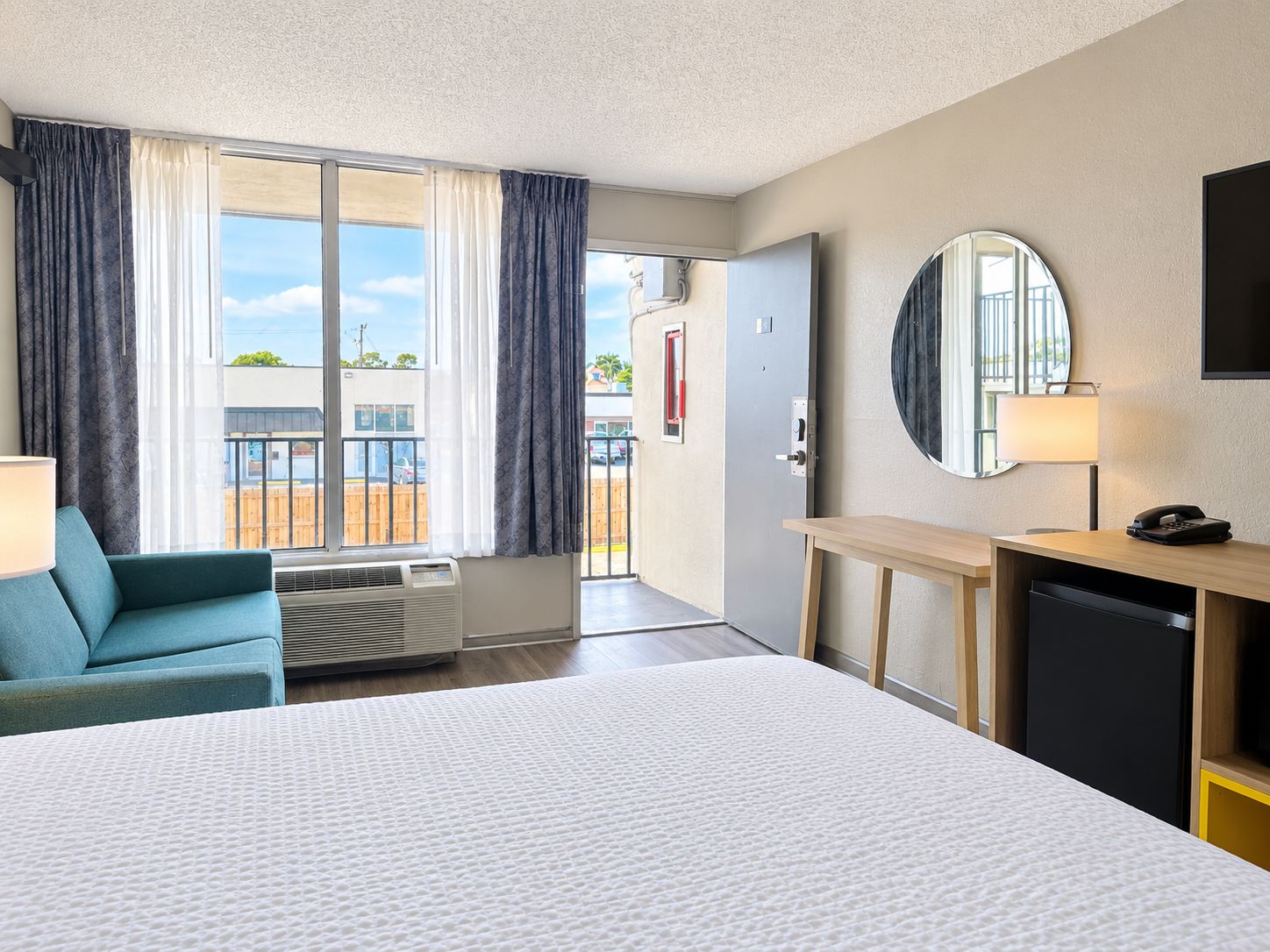 A hotel room with a bed, a desk, a chair, a mirror, a lamp, and a view of the outside.