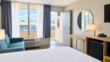 A hotel room with a bed, a desk, a chair, a mirror, a lamp, and a view of the outside.