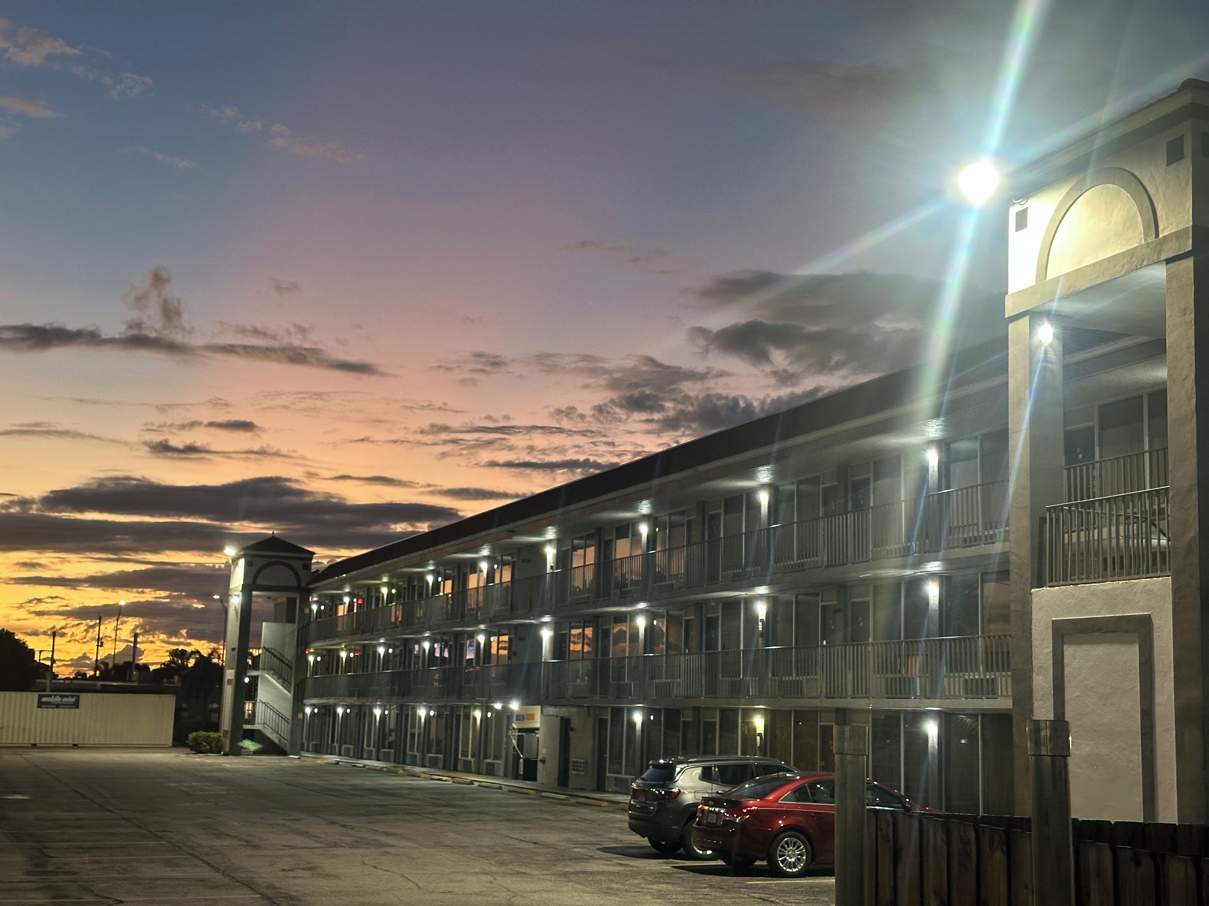 A multi-story hotel with a row of rooms, a parking area, and a sunset sky.
