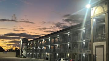 A multi-story hotel with a row of rooms, a parking area, and a sunset sky.