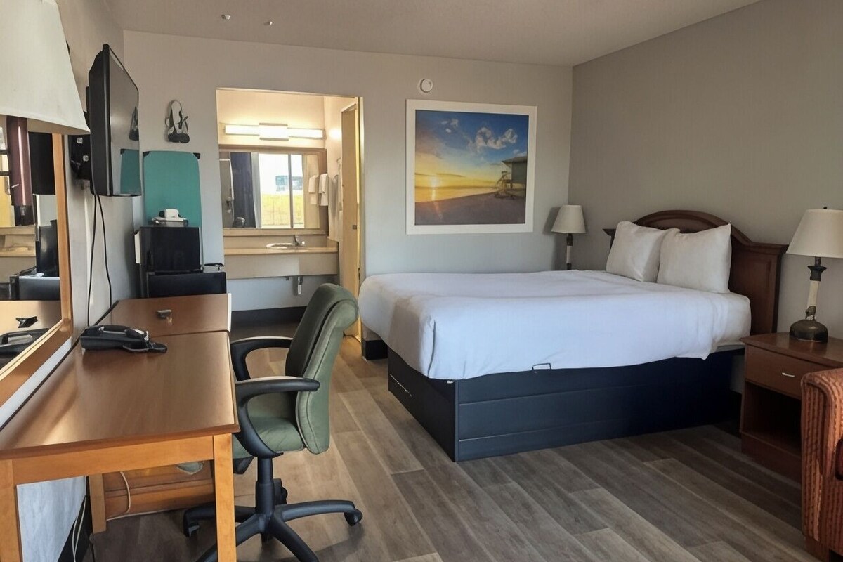A hotel room with a bed, desk, chair, and a painting on the wall.
