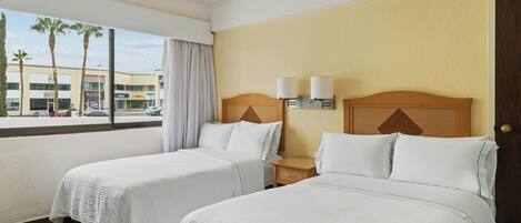 Habitación de hotel con dos camas, una ventana que ofrece vistas a palmeras y un edificio, y un cabecero de madera con una lámpara.