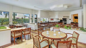 Un vaste espace dédié à la salle à manger, avec de nombreuses tables et chaises, de grandes fenêtres donnant sur un paysage verdoyant et, au fond, une cuisine moderne.