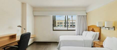 A hotel room with two beds, a desk, a chair, a window with a view of buildings, and a lamp on the wall.