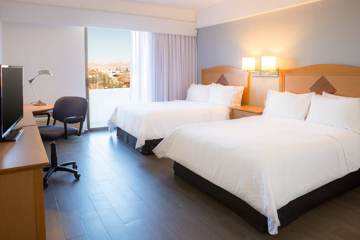 Habitación de hotel con dos camas, un escritorio con computadora, una silla y vista a la ciudad.
