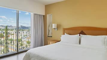 A hotel room with a large bed, a nightstand, a lamp, and a view of a cityscape and mountains through a large window.