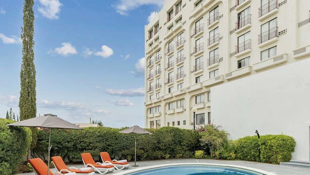 Une piscine entourée de chaises longues et de parasols, avec une végétation luxuriante et un grand immeuble en arrière-plan.
